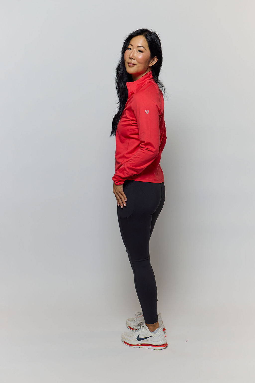 A woman stands in profile wearing a bright red athletic jacket, black leggings, and white Nike running shoes against a plain light gray studio background.