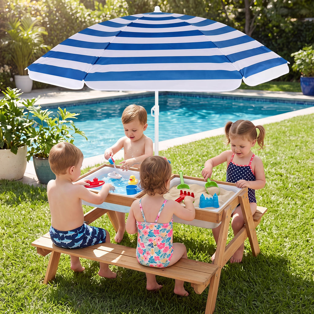 A children's outdoor sand play table set on a grassy lawn next to a swimming pool, with four young children playing under a blue and white striped patio umbrella. The table has a light wood frame and bench, a white sand tray, and colorful toy accessories.