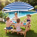 A children's outdoor sand play table set on a grassy lawn next to a swimming pool, with four young children playing under a blue and white striped patio umbrella. The table has a light wood frame and bench, a white sand tray, and colorful toy accessories.