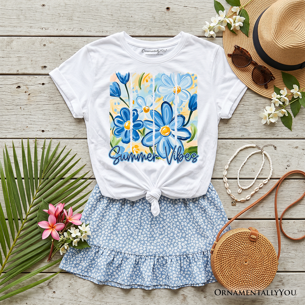 White t-shirt with blue floral design and 'Summer Vibes' text, paired with a blue floral skirt, straw hat, sunglasses, and round bag on a wooden surface.