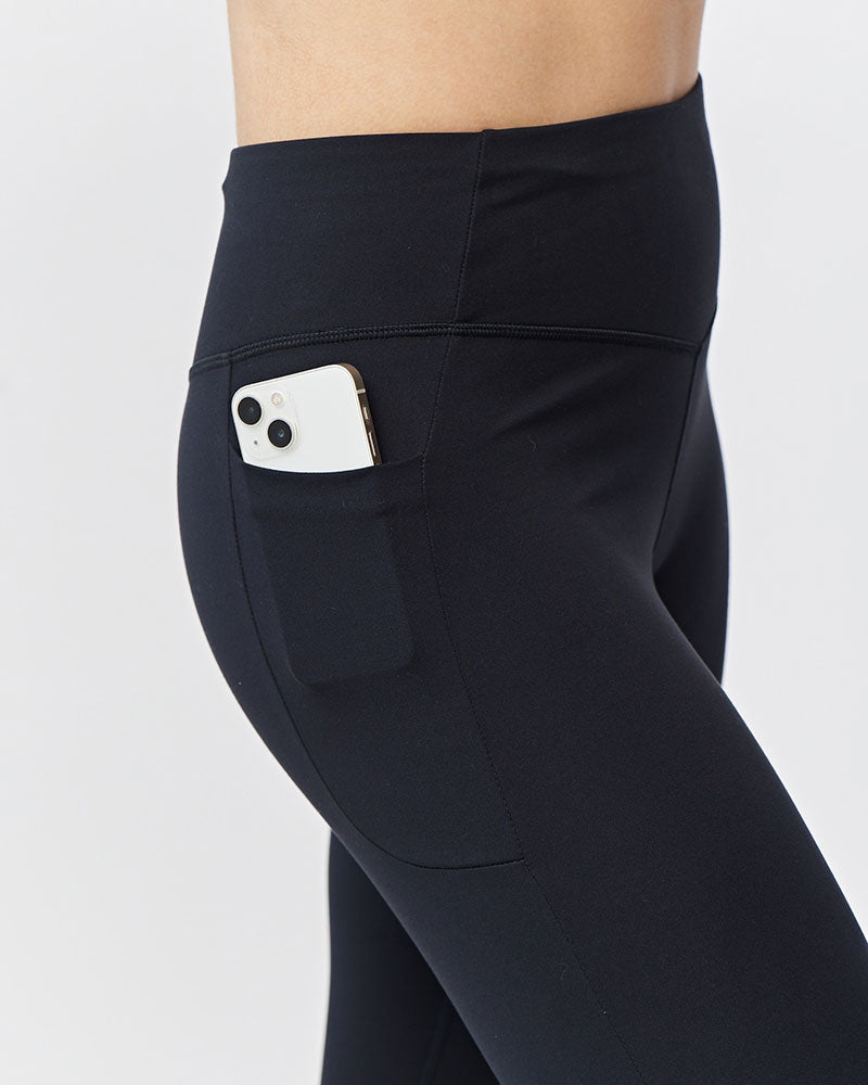 Close-up side view of a person wearing black high-waisted leggings with a side pocket holding a white smartphone, against a light gray studio background.