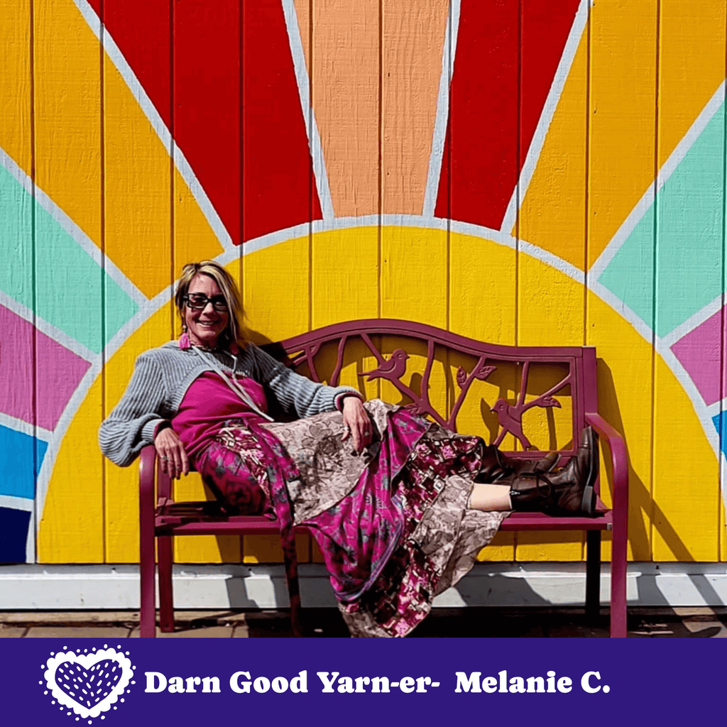 Woman sitting on a bench in front of a colorful wooden wall with 'Darn Good Yarn-er' branding.