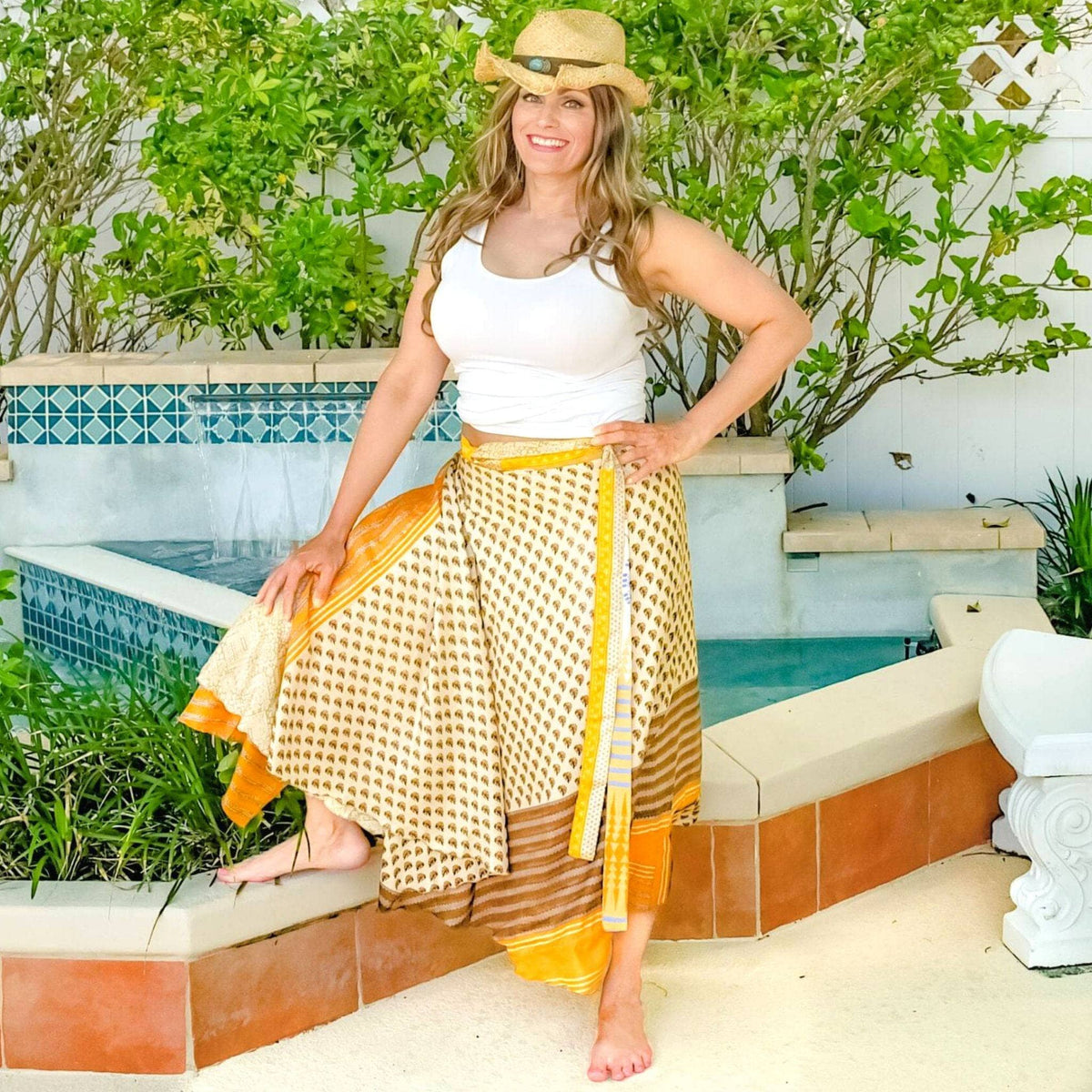 Woman in a white top and yellow patterned skirt standing by a pool with greenery in the background