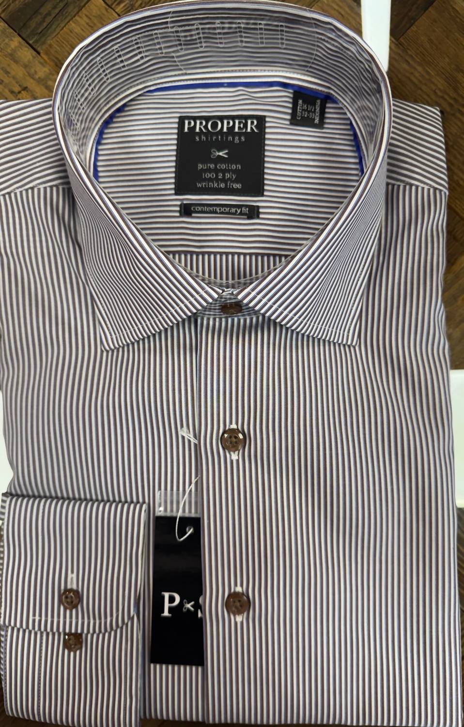 A folded long-sleeve dress shirt by Proper Stitchers, featuring a fine vertical stripe pattern in navy blue and white. The shirt has a classic collar, button-down front, and a visible hangtag.